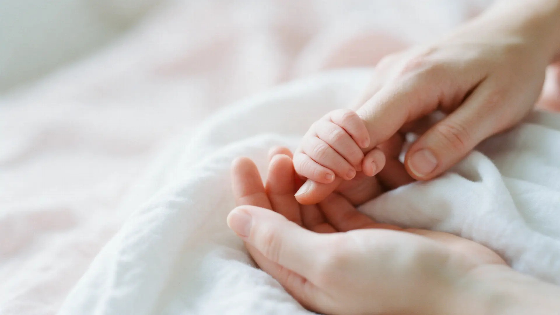 A newborn's tiny hand holding a parent's finger on a soft white blanket