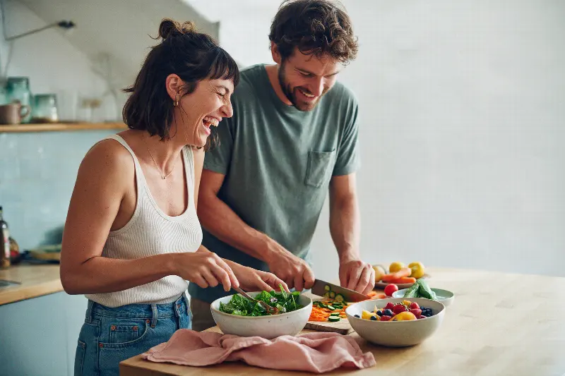 A couple laughing while they make a salad together in a bright kitchen