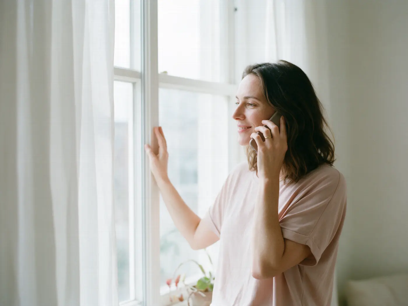 A woman by a bright window smiling as she talks on the phone