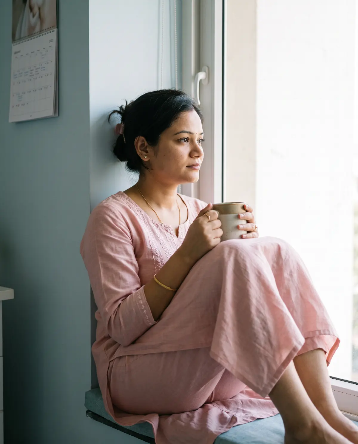 A woman on a window seat with a warm mug, gazing out with a quiet, hopeful look