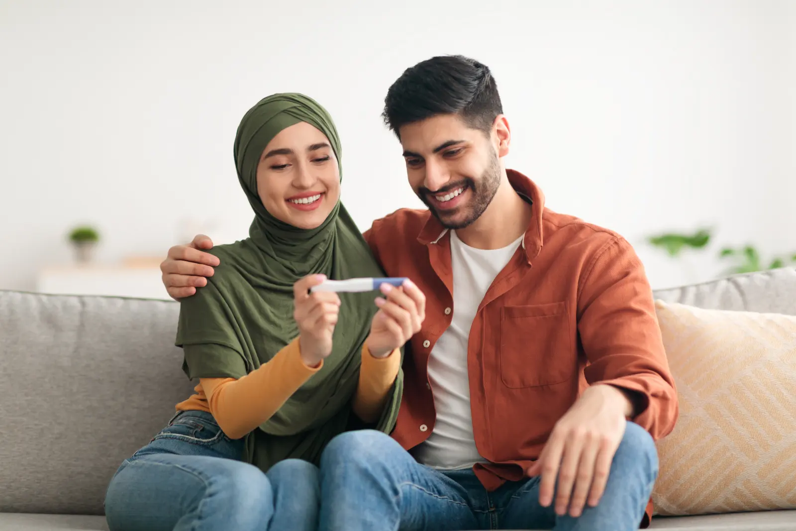 A couple laughing as they hold a positive pregnancy test on their sofa