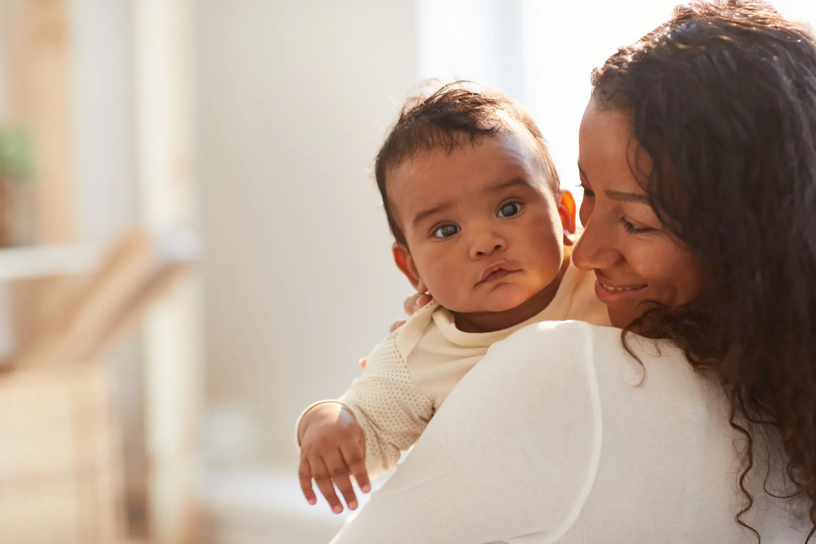 A mother smiling as she holds her baby close by a bright window