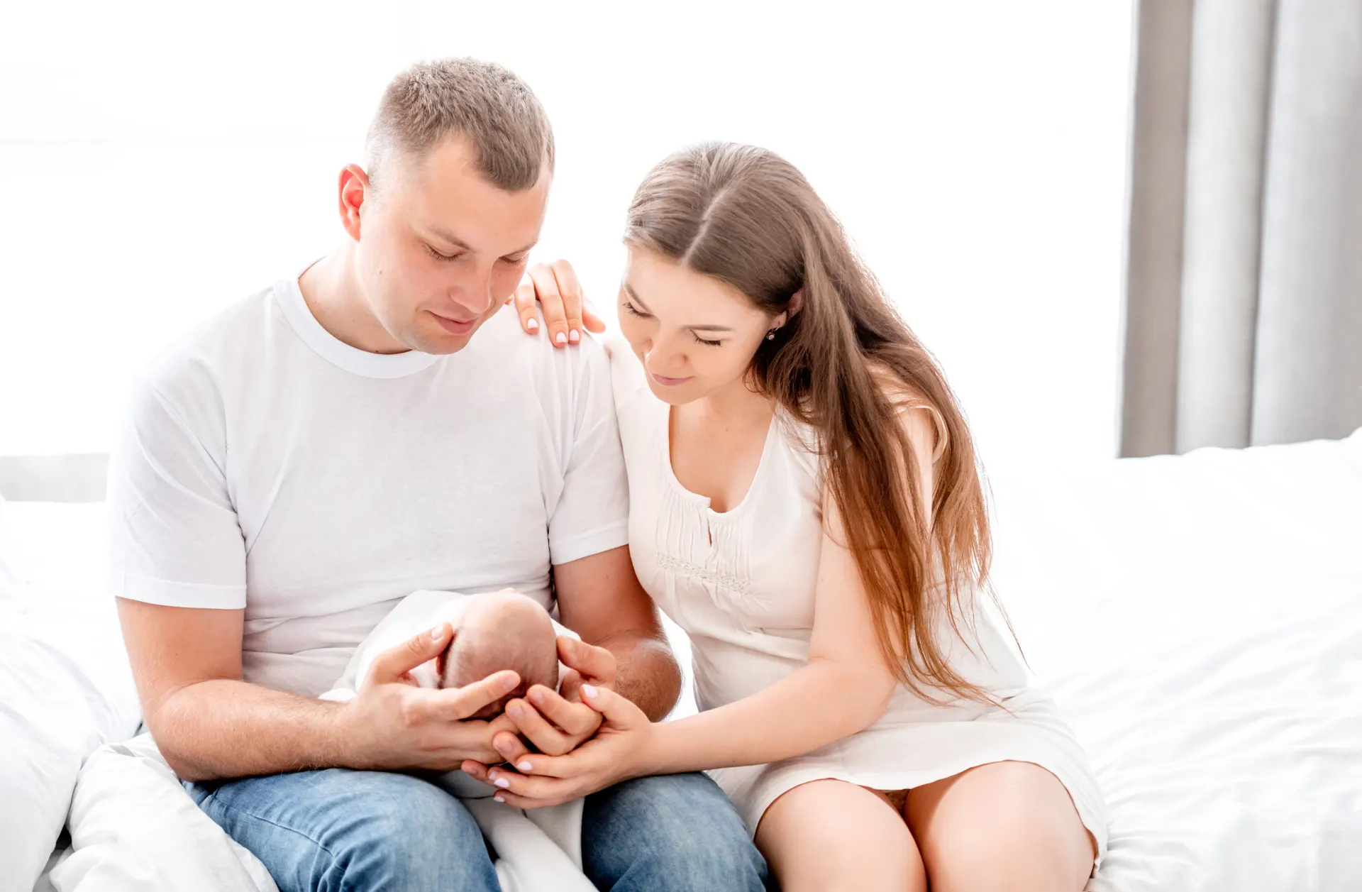 New parents sitting on a bed, holding their sleeping newborn in morning light