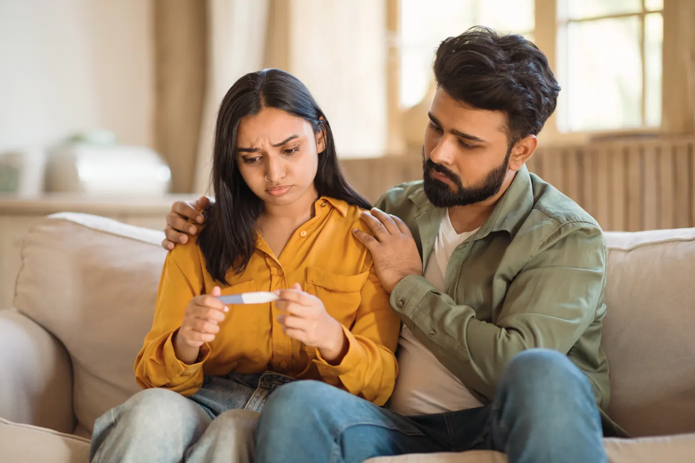 A young couple on a sofa looking at a pregnancy test, his arm around her shoulders