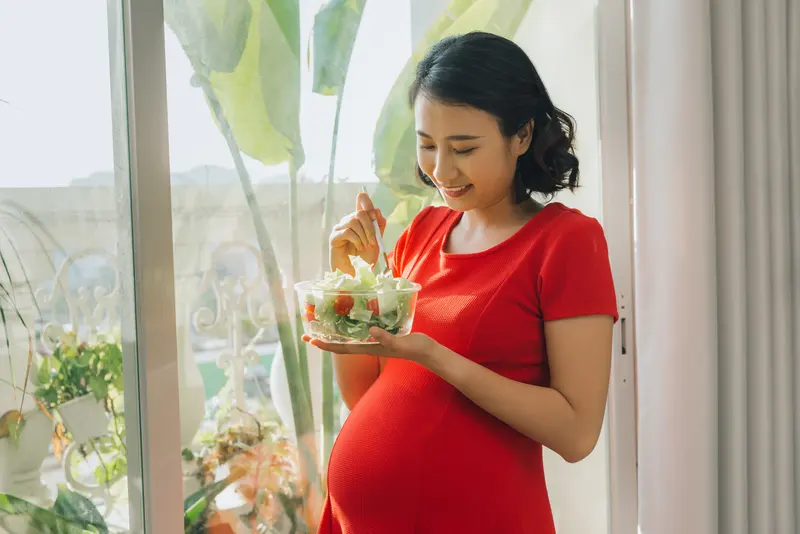 A smiling pregnant woman eating a bowl of fresh salad by a bright window