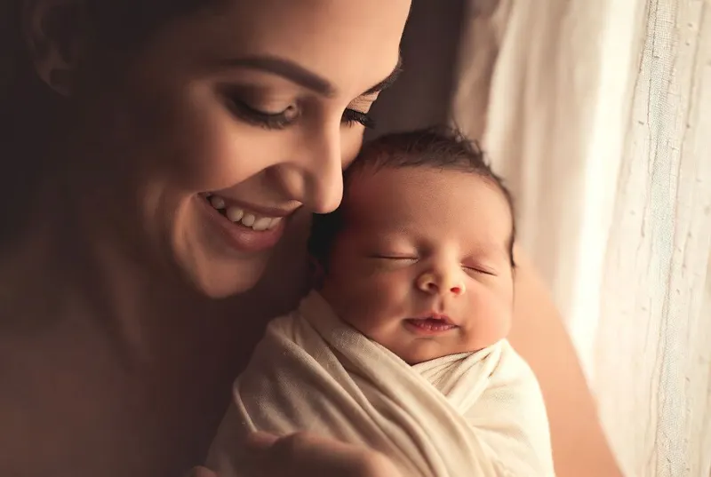 A smiling mother holding her sleeping newborn close in soft light